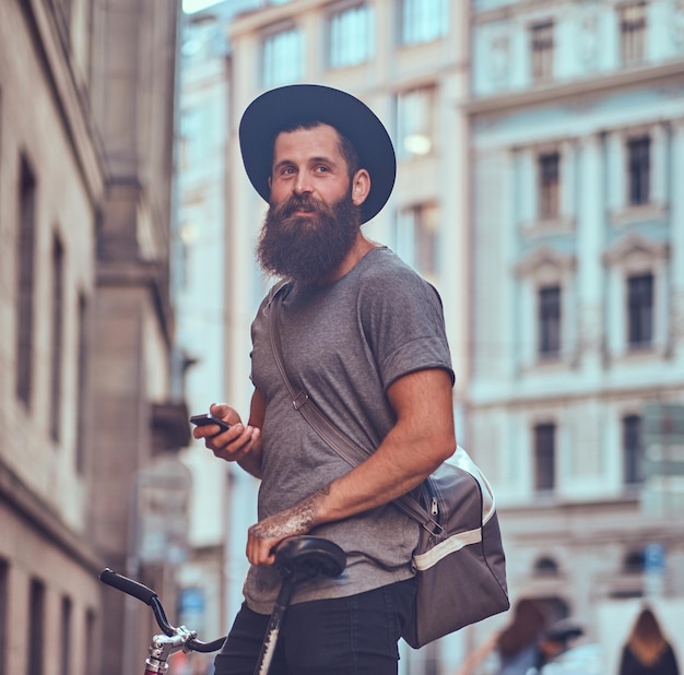 Bel viaggiatore hipster con una barba elegante e un tatuaggio sulle braccia vestito con abiti casual con una borsa, si trova sul marciapiede, riposando dopo un giro in bicicletta.