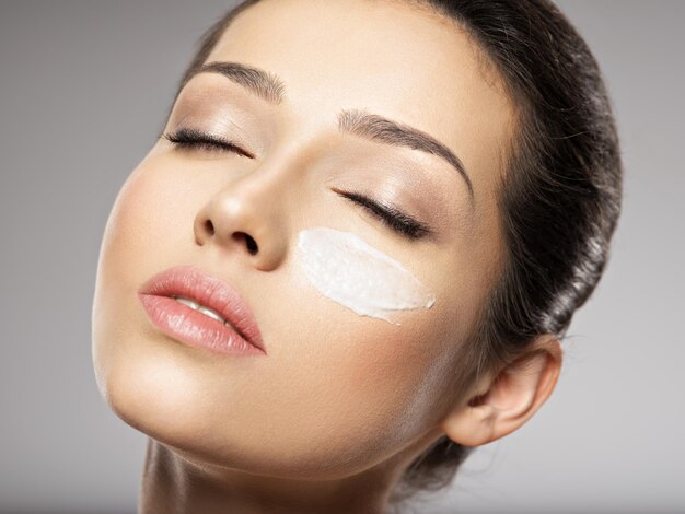 Bel viso di giovane donna con crema cosmetica striscio sul viso vicino all'occhio. Concetto di cura della pelle. Concetto di trattamento di bellezza.