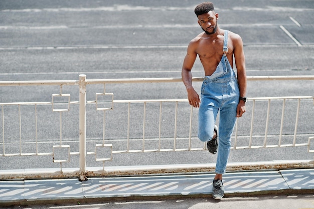 Bell'uomo a torso nudo afroamericano sexy in tuta di jeans posato allo stadio Ritratto di uomo nero alla moda