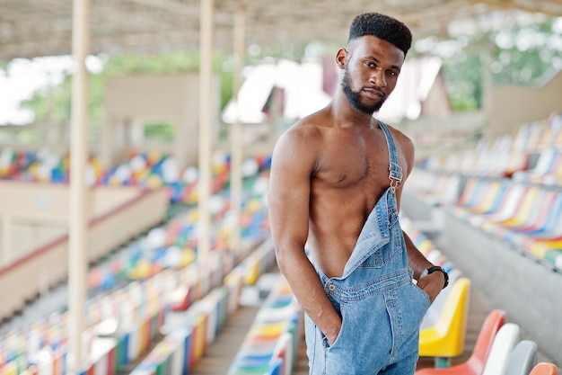 Bell'uomo a torso nudo afroamericano sexy in tuta di jeans posato su sedie colorate allo stadio Ritratto di uomo nero alla moda