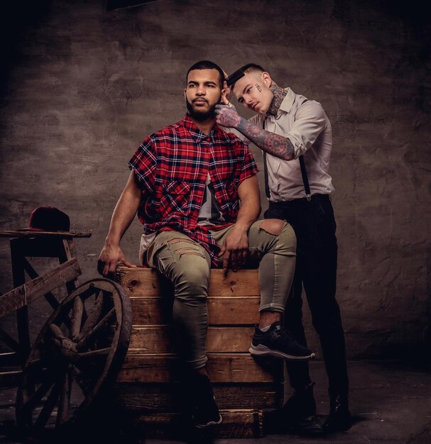 Bell'uomo afroamericano che si taglia i capelli mentre è seduto su scatole di legno in uno studio. Il parrucchiere professionista tatuato vecchio stile si taglia i capelli.