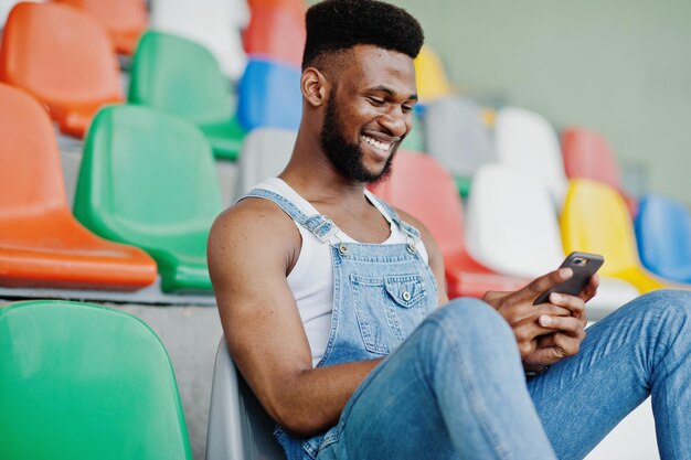 Bell'uomo afroamericano in tuta di jeans con telefono cellulare a portata di mano poste su sedie colorate allo stadio Ritratto di uomo nero alla moda