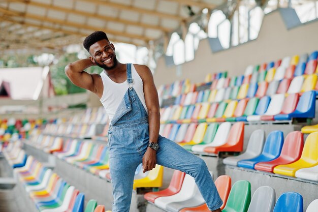 Bell'uomo afroamericano in tuta di jeans posata su sedie colorate allo stadio Ritratto di uomo nero alla moda