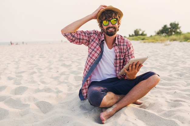 Bell'uomo allegro con la barba in cappello di paglia, camicia a scacchi e occhiali da sole alla moda che si siede sulla sabbia bianca e utilizzando tablet