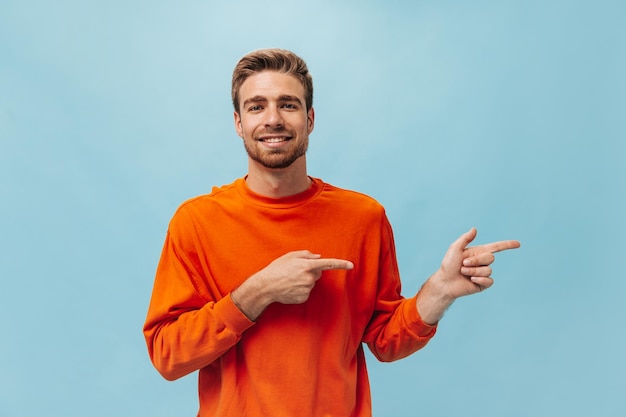 Bell'uomo barbuto con acconciatura alla moda in felpa arancione brillante che guarda nella fotocamera sorridente e indica il posto per il testo