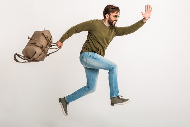 Bell'uomo barbuto elegante che salta in esecuzione isolato vestito in felpa con borsa da viaggio, indossa jeans e occhiali da sole, viaggiatore pazzo in fretta