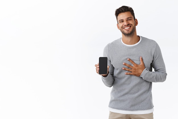 Bell'uomo barbuto maschile contento e lusingato in maglione grigio che mostra i suoi risultati di fitness sull'app mobile che tocca il petto grato o orgoglioso sorridente con gioia promuovere l'applicazione per smartphone