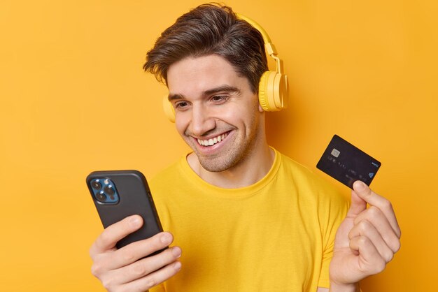 Bell'uomo brunet guarda felicemente lo smartphone controlla il suo conto bancario effettua il pagamento online tramite l'applicazione ascolta la traccia audio tramite le cuffie tiene la carta di credito isolata su sfondo giallo