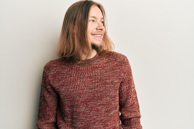 Bell'uomo caucasico con i capelli lunghi che indossa un maglione invernale casual che guarda di lato, rilassa la posa del profilo con il viso naturale e il sorriso sicuro.