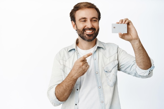 Bell'uomo caucasico sorridente che dimostra la nuova carta di credito che sembra soddisfatto in piedi su sfondo bianco Spazio di copia