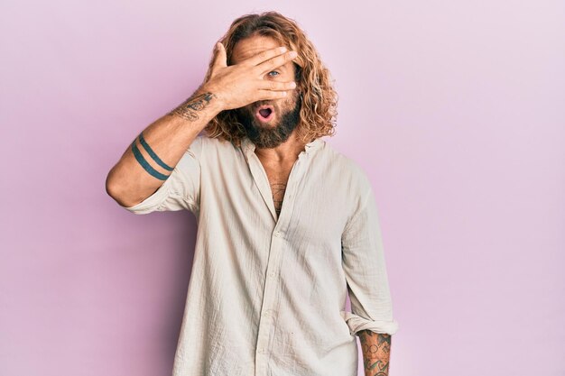 Bell'uomo con barba e capelli lunghi che indossa abiti casual che sbirciano sotto shock coprendo viso e occhi con la mano che guarda attraverso le dita impaurite