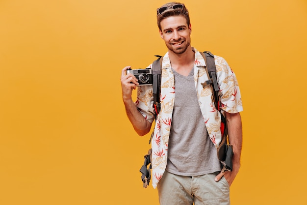 Bell'uomo con la barba allo zenzero in camicia bianca e t-shirt scozzese che sorride e tiene la macchina fotografica sul muro arancione isolato