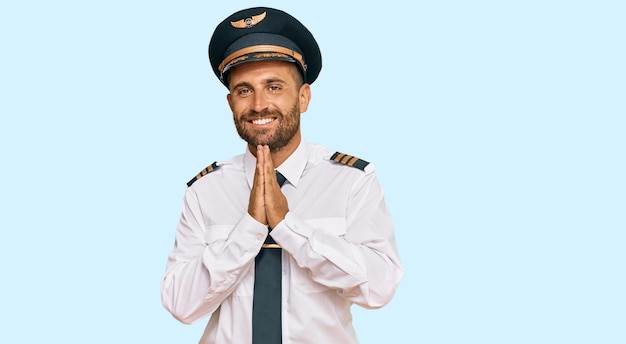 Bell'uomo con la barba che indossa l'uniforme da pilota di aeroplano che prega con le mani insieme chiedendo perdono sorridendo fiducioso.