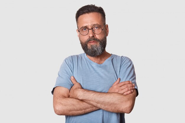 bell'uomo con la barba in piedi con le braccia conserte isolato su bianco, sembra serio, indossa maglietta grigia casual e occhiali, posa maschile. Concetto di persone