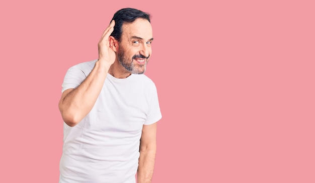 Bell'uomo di mezza età che indossa una maglietta casual sorridente con la mano sull'orecchio che ascolta un'audizione di voci o pettegolezzi. concetto di sordità.
