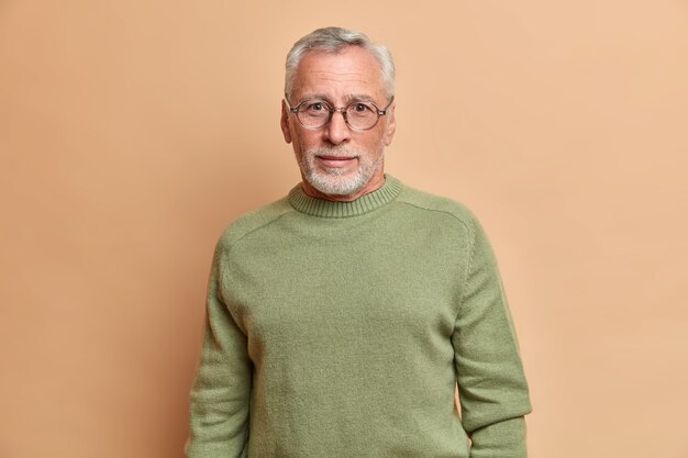 Bell'uomo europeo barbuto con lo sguardo curioso indossa occhiali e il maglione di base guarda direttamente in pose frontali contro il muro beige