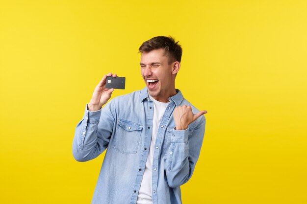 Bell'uomo felice che celebra la vittoria, trionfando. Ragazzo che mostra la carta di credito e grida sì dallo stupore e dalla gioia, riceve bonus, cashback extra, sfondo giallo in piedi.