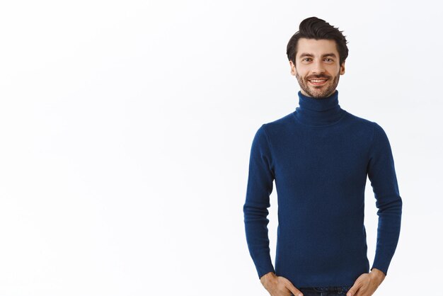 Bell'uomo fiducioso con setole in elegante maglione blu a collo alto tenere le mani nelle tasche dei jeans sorridendo con gioia aspetto assertivo preparandosi per la festa di capodanno stand sfondo bianco