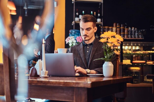 Bell'uomo freelance con barba e capelli alla moda vestito con un abito nero che lavora su un laptop mentre è seduto in un caffè.