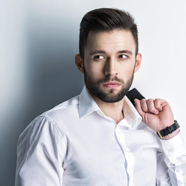 Bell'uomo in camicia bianca tiene il vestito nero - posa sul muro. Ragazzo attraente con acconciatura di moda. Uomo fiducioso con barba corta. Ragazzo adulto con capelli castani.