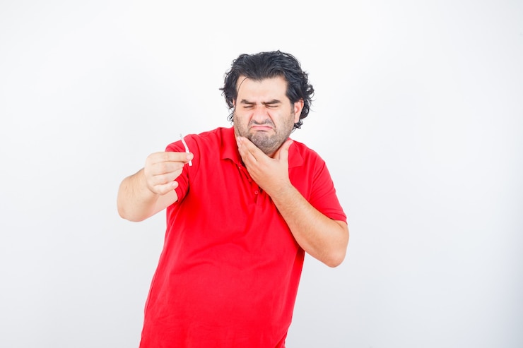 Foto gratuito bell'uomo in maglietta rossa che tiene sigaretta, tenendo la mano sul collo, facendo smorfie e guardando dispiaciuto, vista frontale.