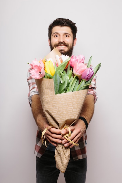 Bell'uomo innamorato che desidera felice giorno di San Valentino, dando bouquet di fiori in un appuntamento romantico, sorridente, vestito da portare sopra il muro bianco