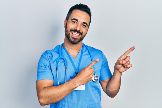 Bell'uomo ispanico con la barba che indossa l'uniforme del medico sorridendo e guardando la telecamera che punta con due mani e le dita di lato.