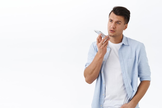 Bell'uomo macho dall'aspetto serio in camicia registra un messaggio vocale o un memo nell'app per smartphone che tiene il telefono cellulare vicino alle labbra vicino alla conversazione dinamica con lo sfondo bianco dell'assistente di persona