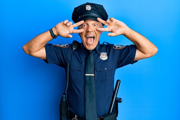 Bell'uomo maturo di mezza età che indossa l'uniforme della polizia che fa il simbolo della pace con le dita sul viso, sorridendo allegro mostrando vittoria