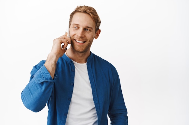 Bell'uomo moderno dai capelli rossi che parla al telefono, chiama un amico e sorride, tiene lo smartphone vicino all'orecchio e guarda da parte, in piedi su sfondo bianco