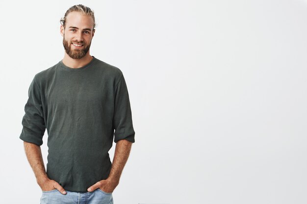 Bell'uomo nordico con barba e acconciatura elegante in camicia grigia e jeans sorridenti, tiene le mani in tasca.