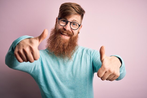 Bell'uomo rosso irlandese con la barba che indossa occhiali su sfondo rosa isolato che approva facendo un gesto positivo con il pollice in alto sorridente e felice per il successo Gesto del vincitore