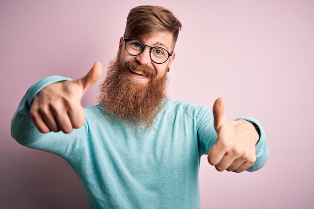 Bell'uomo rosso irlandese con la barba che indossa occhiali su sfondo rosa isolato che approva facendo un gesto positivo con il pollice in alto sorridente e felice per il successo Gesto del vincitore