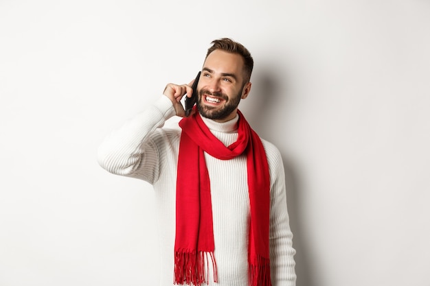Bell'uomo sorridente che parla al telefono, sembra soddisfatto, in piedi con sciarpa e maglione invernali, sfondo bianco