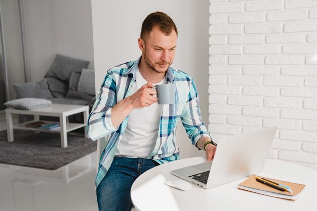 Bell'uomo sorridente in camicia seduto in cucina a casa al tavolo che lavora online sul laptop da casa