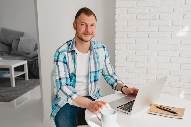 Bell'uomo sorridente in camicia seduto in cucina a casa al tavolo che lavora online sul laptop da casa