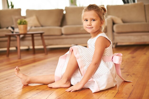 Bella affascinante bambina che indossa un abito festivo con gonna ampia seduta a piedi nudi sul pavimento della cucina