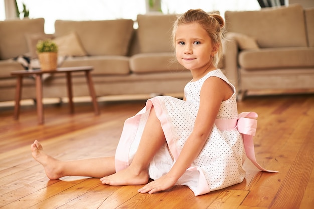 Bella affascinante bambina che indossa un abito festivo con gonna ampia seduta a piedi nudi sul pavimento della cucina