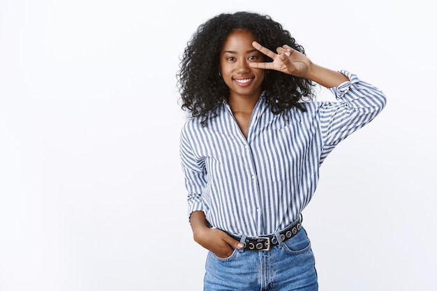 Bella affascinante sorridente felice giovane ragazza dalla pelle scura dai capelli ricci che indossa jeans blusa a righe alla moda che mostra il segno della vittoria della pace della discoteca sull'occhio che sorride con gioia in posa carino colpo muro bianco