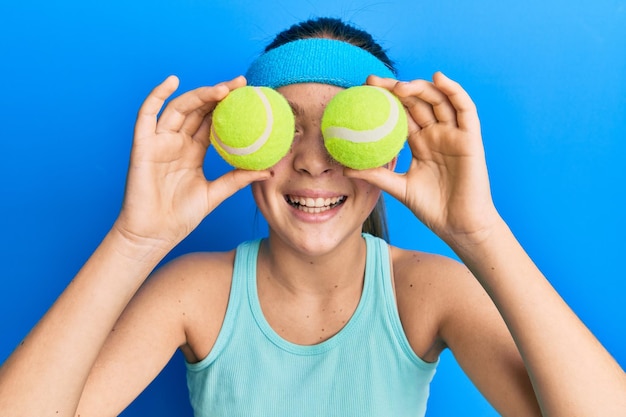 Bella bambina castana che tiene la pallina da tennis vicino agli occhi sorridendo e ridendo ad alta voce perché divertente scherzo pazzo.