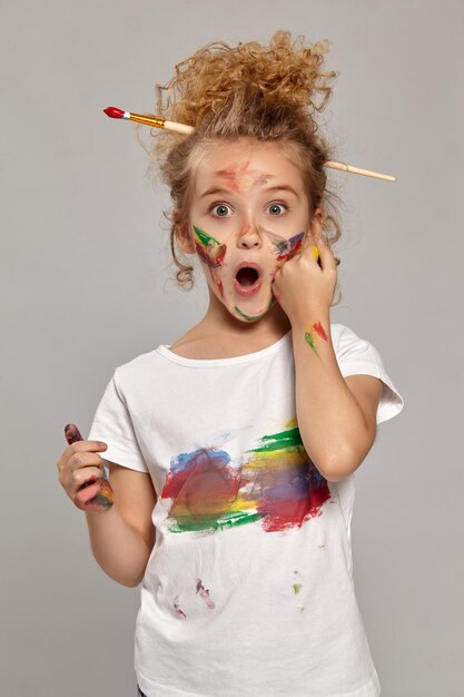 Bella bambina che ha una spazzola tra i suoi capelli biondi ricci chic, indossa una maglietta bianca, con le dita dipinte, sembra meravigliata su uno sfondo grigio.