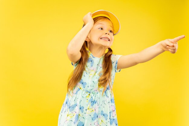 Bella bambina emotiva isolata su sfondo giallo. Ritratto a mezzo busto di bambino felice che indossa un vestito e berretto arancione rivolto verso l'alto. Concetto di estate, emozioni umane, infanzia.