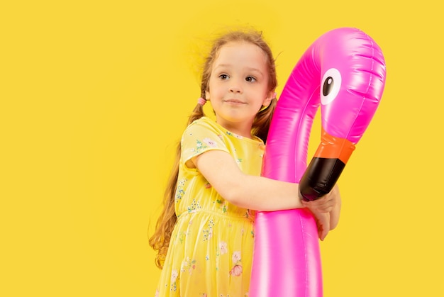 Bella bambina emotiva isolata su sfondo giallo. Ritratto a mezzo busto di bambino felice che indossa un vestito e che tiene il fenicottero rosa di gomma. Concetto di estate, emozioni umane, infanzia.