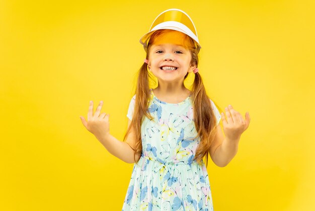 Bella bambina emotiva isolata su sfondo giallo. Ritratto a mezzo busto di bambino felice e in festa che indossa un vestito e un berretto arancione. Concetto di estate, emozioni umane, infanzia.