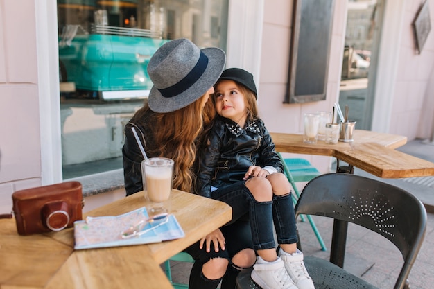 Bella bambina in jeans alla moda e giacca nera che si siede sulle ginocchia della madre e la guarda con amore.
