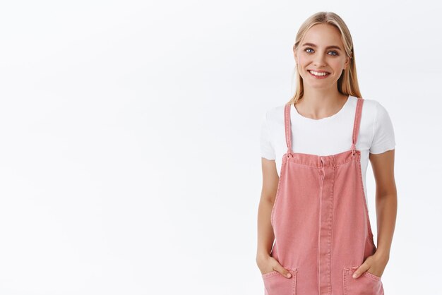 Bella bella donna bionda femminile in salopette rosa maglietta tenere le mani in tasca in posa rilassata casual sorridente con gioia esprimere atteggiamento amichevole e positivo sfondo bianco