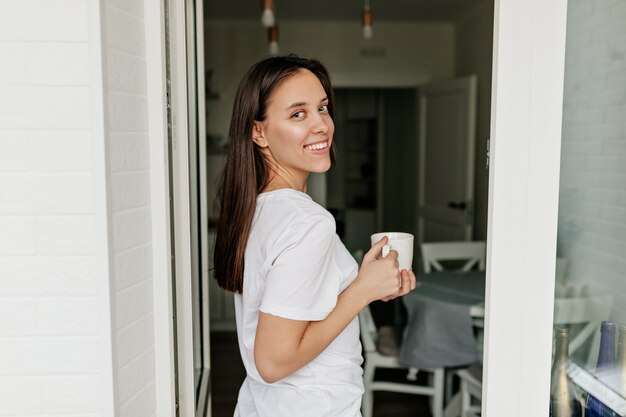bella bella donna con i capelli scuri, sorriso felice che indossa una maglietta bianca che beve il caffè del mattino in cucina. buona giornata di sole.