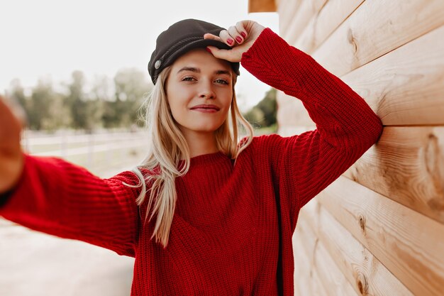 Bella bionda che fa selfie vicino al legno. Donna attraente in vestiti rossi alla moda.