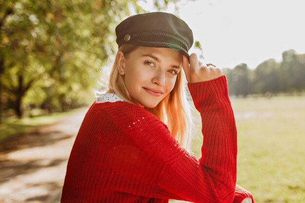 Bella bionda che sembra carina in pullover rosso e cappello nero alla moda in piedi nel parco. La ragazza sorridente gode di una giornata di sole autunnale.