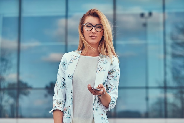 Bella blogger alla moda in abiti eleganti e occhiali con una borsetta, tiene uno smartphone, in piedi contro un grattacielo.
